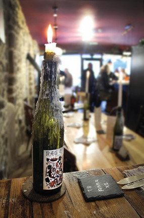 A candle burns atop a wax-covered wine bottle on a rustic table in a cozy, dimly-lit room with people in the blurred background.