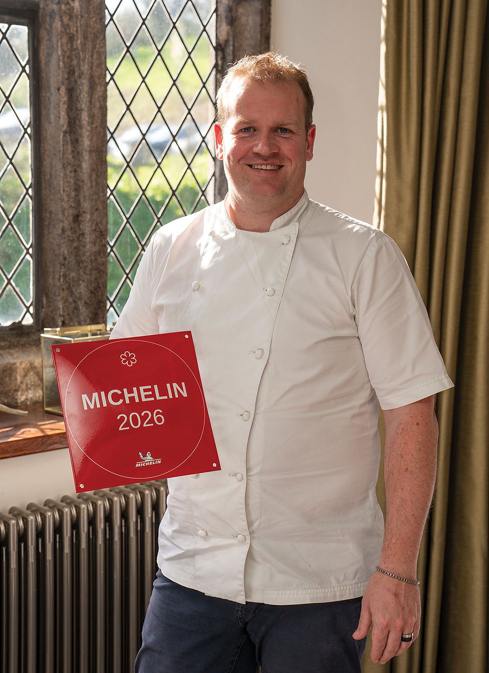 Chef in white uniform holds a red "MICHELIN 2026" sign, smiling beside a window with diamond patterns. Sunny and cheerful ambiance.