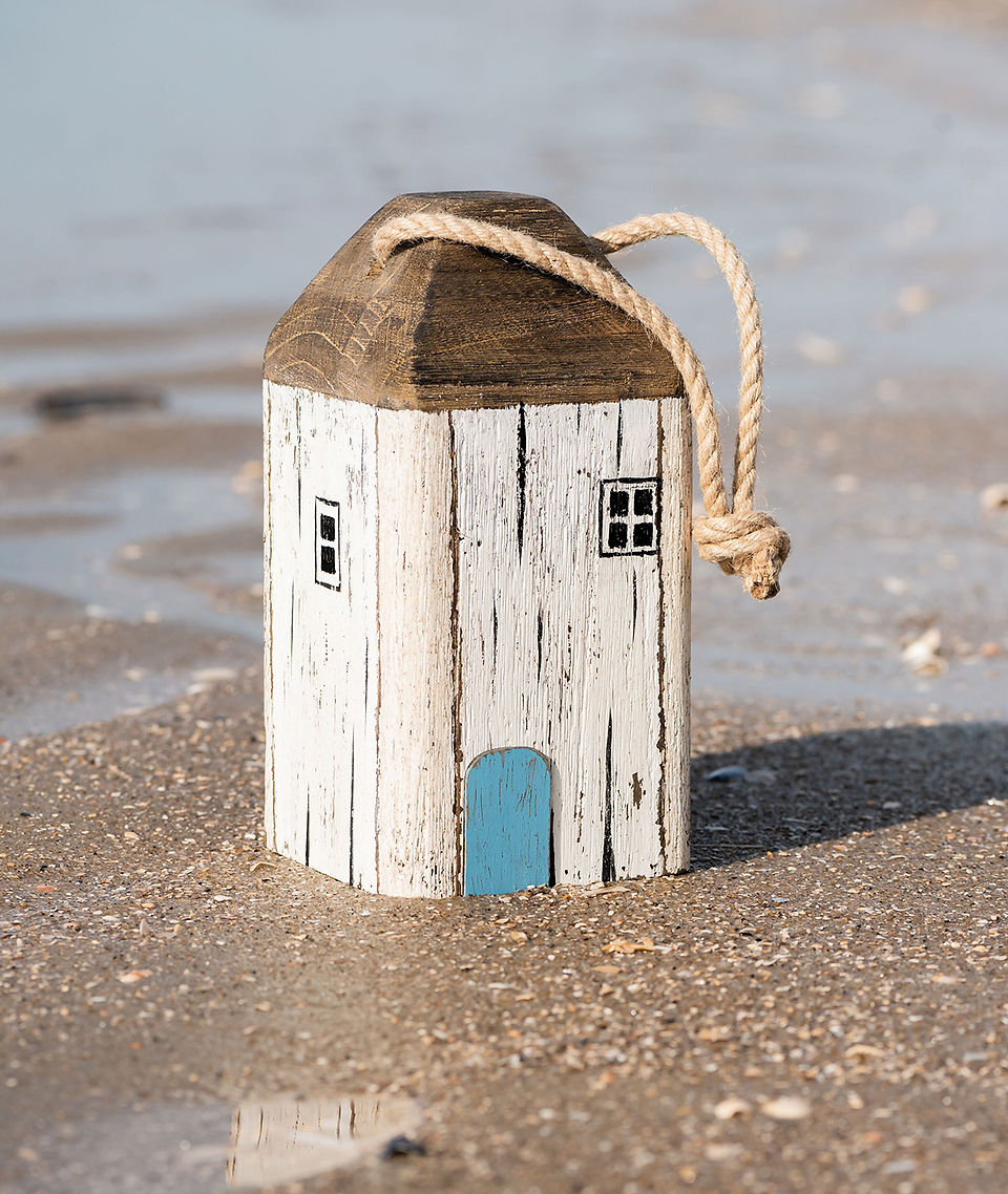 Painted wooden house ornament with rope handle on sandy beach, blue door, calm seaside backdrop.
