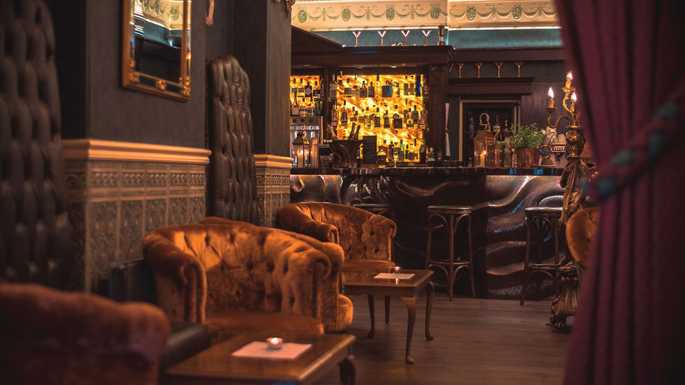 Cozy lounge with vintage velvet couches, dim lighting, ornate decor, and a glowing bar in the background. Luxurious and inviting ambiance.