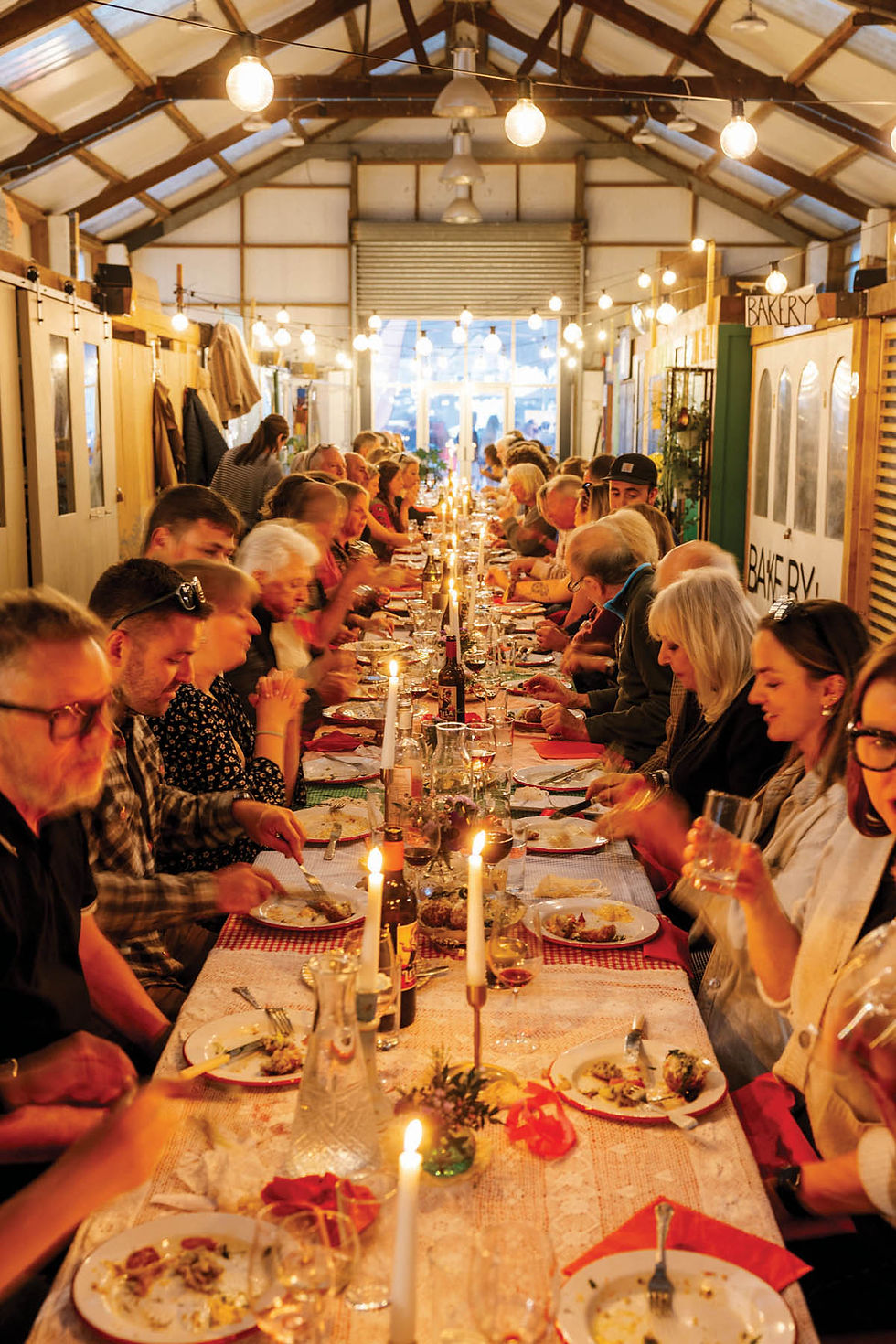 A long, candlelit dinner table filled with people eating and talking in a cozy, warmly lit room. Signs reading "Bakery" visible on walls.