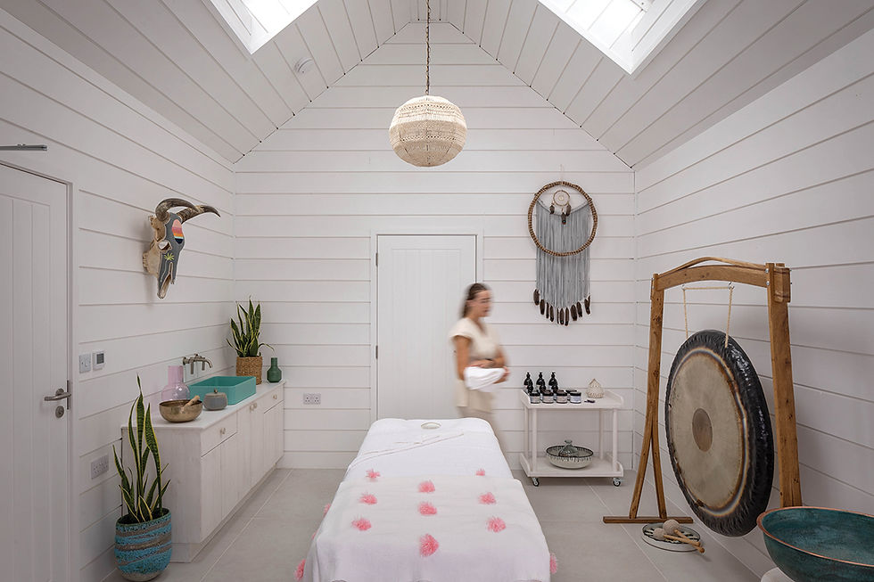 the relaxing massage and treatment room at Three Mile Beach in Hayle. Bright white spa room with a blurred woman walking past a massage bed, gong, dreamcatcher, and teal bowls.