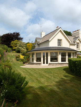 Cream-colored cottage with round conservatory in a lush green garden under a cloudy blue sky.
