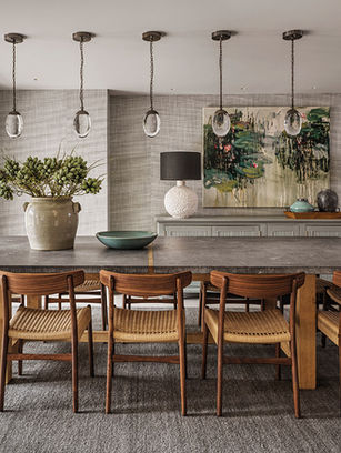 Modern dining room with long stone table, wooden chairs, pendant lights, abstract wall art, and lamps on a sideboard