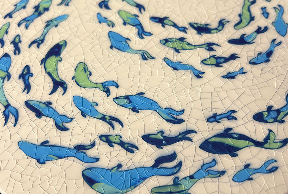Round white ceramic plate with blue and green fish or dolphin shapes in a swirling mosaic pattern, viewed from above.