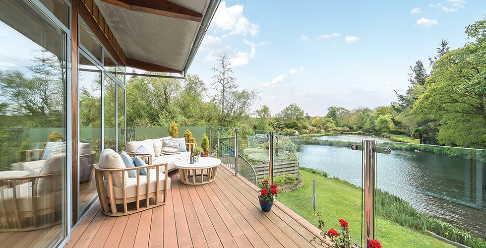 Spacious deck with cozy furniture overlooking a serene lake. Glass railing, lush greenery, red potted flowers, and a clear blue sky.