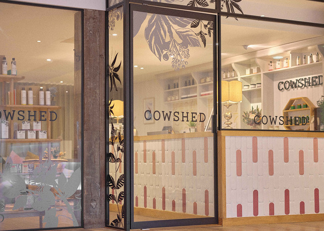 Glass storefront of a spa labeled "Cowshed" with leaf designs. Interior shows products, a lit lamp, and warm tones, giving a cozy vibe.