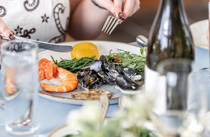 Person dining with seafood platter, including shrimp and mussels, on a blue tablecloth. A hand holds a fork and knife.