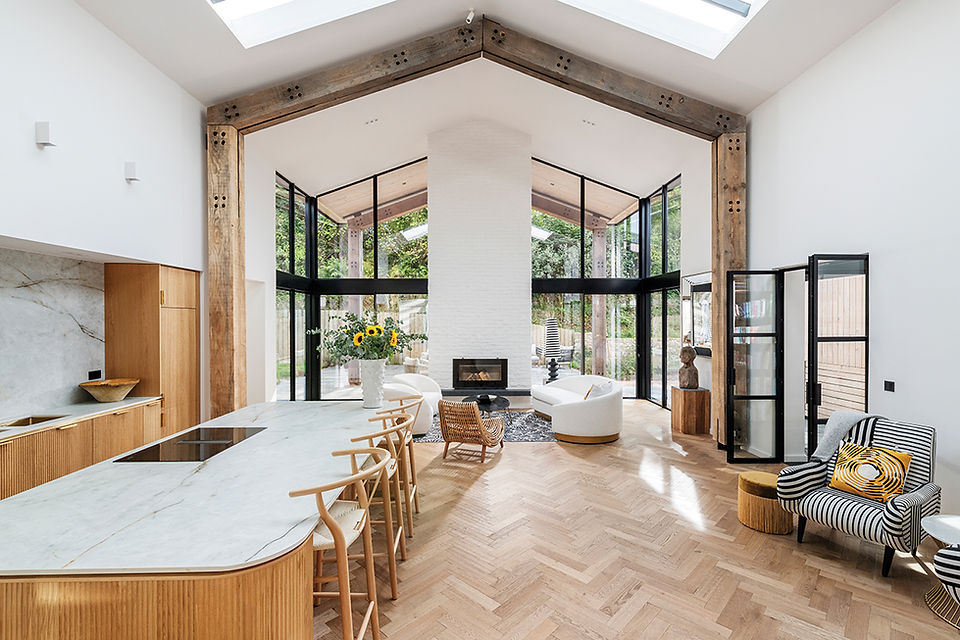 Bright modern living room with vaulted skylights, wood beams, glass walls, fireplace, marble island, and striped chair.