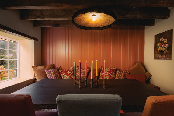 Cozy dining nook with a dark table, colorful candles, patterned cushions, wood beams, and a warm orange paneled wall.