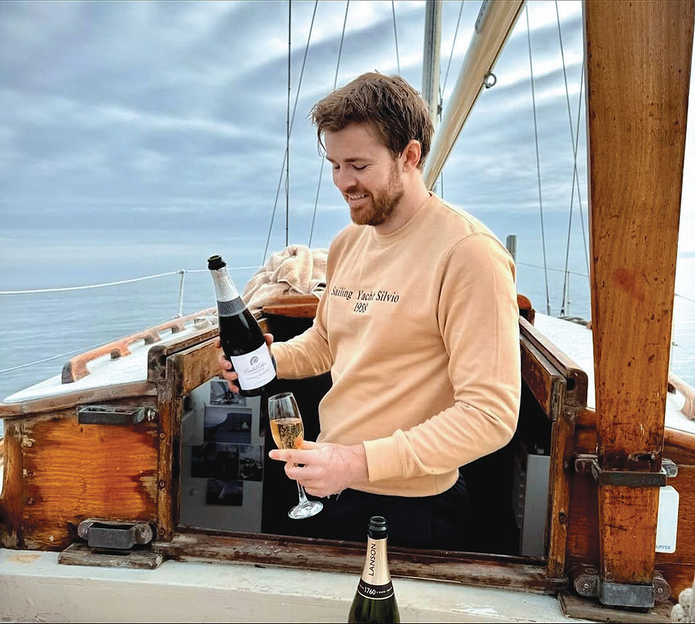 Man on a sailboat pours champagne into a flute on calm water under cloudy sky, wearing Sailing Yacht Silvio 1988 sweatshirt.