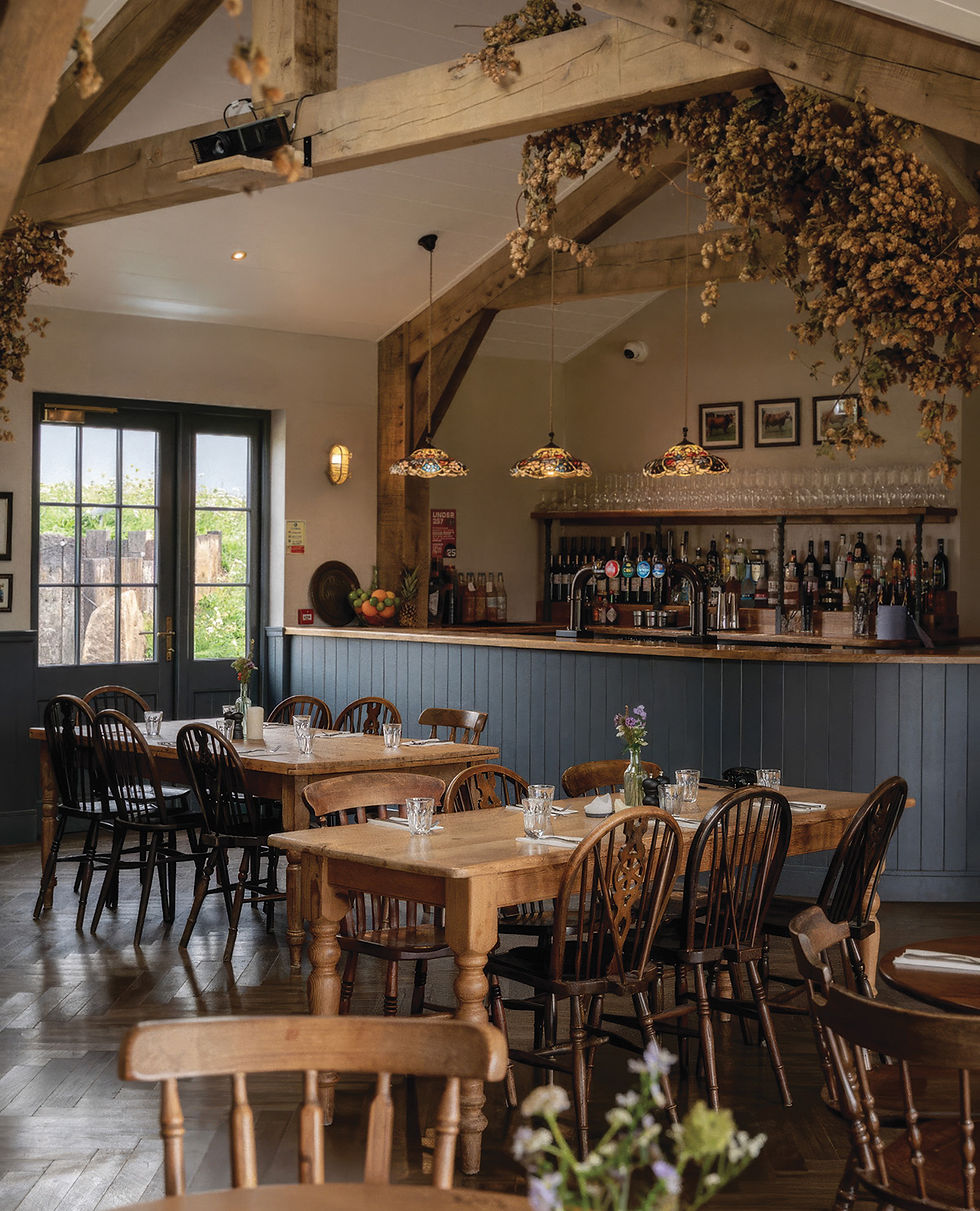Halwyn of Crantock's newly designed Cozy pub interior with wooden beams, rustic tables, and chairs. Decor includes hanging dried hops and stained glass lights above the bar. with an Oak beam interior designed by Post and Beam