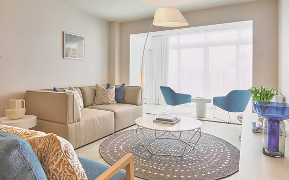 Modern living room with beige sofa, blue chairs, round coffee table on a gray rug. Soft light through large windows, cozy and elegant.