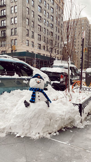 "Snowman with a blue scarf and hat sits atop a snowy mound on a city sidewalk, surrounded by parked cars and tall buildings on a winter day."