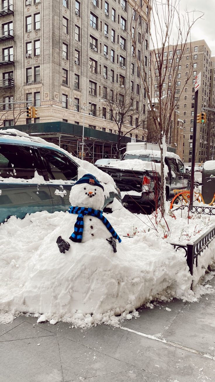 "Snowman with a blue scarf and hat sits atop a snowy mound on a city sidewalk, surrounded by parked cars and tall buildings on a winter day."