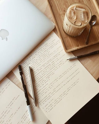 Overhead shot of a white laptop with an Apple logo, a glass of iced coffee with ice cubes on a light wooden tray, papers with handwritten text, two pens (one black and one silver), and a spoon on a light-colored wooden table.
