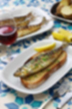 A rustic plate of grilled sardines garnished with lemon slices, paired with a crusty loaf of bread, accompanied by a glass of deep red wine, creating a simple yet elegant Mediterranean feast.