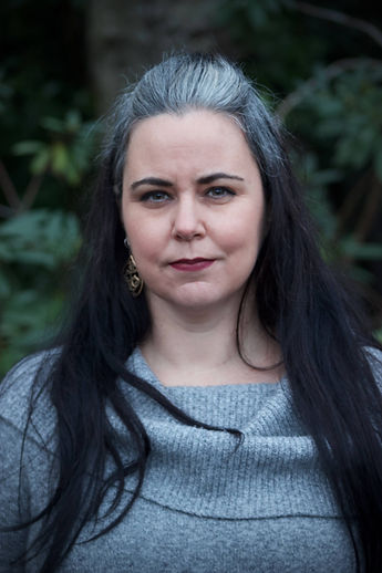 Photo of Elizabeth, a white femme with long salt and pepper hair wearing a grey sweater, trying to smile without looking silly.