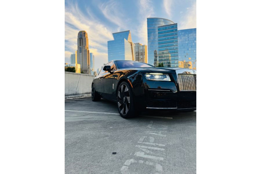 Black Rolls-Royce parked on rooftop with modern city skyline and sunset reflections.