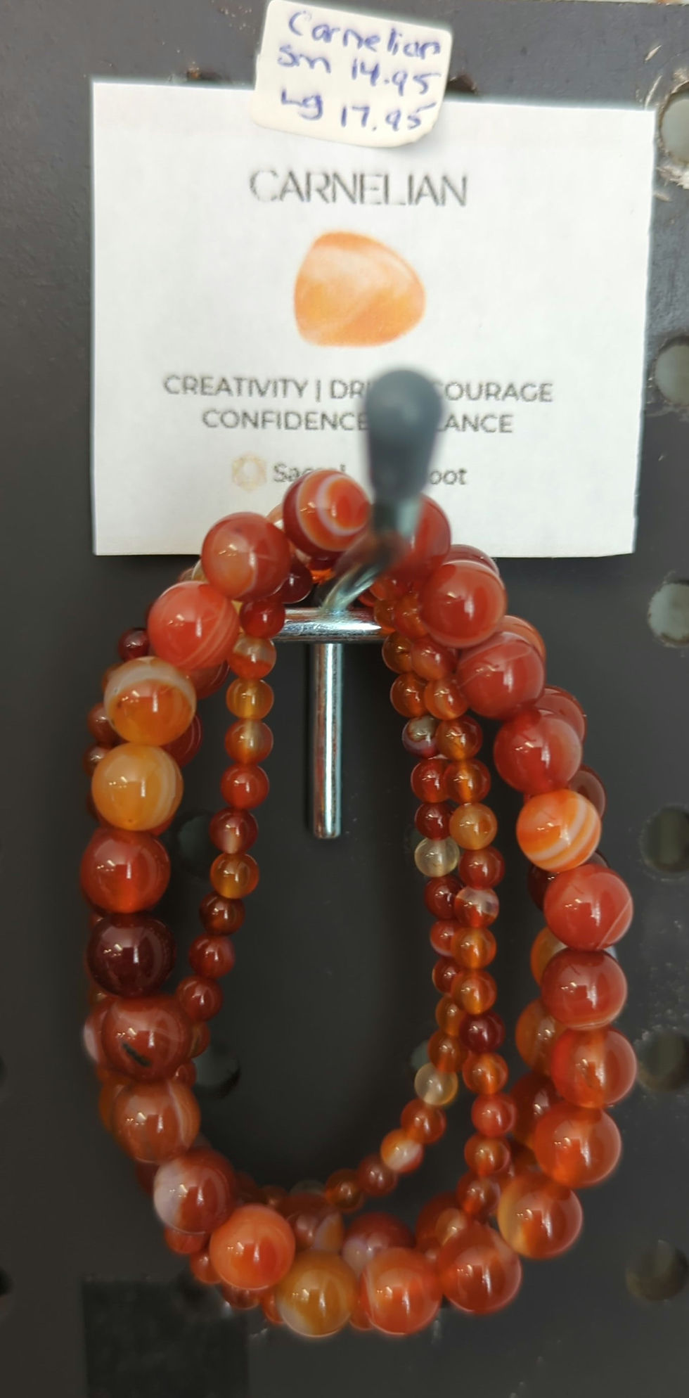 Carnelian bracelets hang on a peg. A label reads "CREATIVITY | DRIVE | COURAGE CONFIDENCE. Small $14.95, Large $17.95" against a dark background.