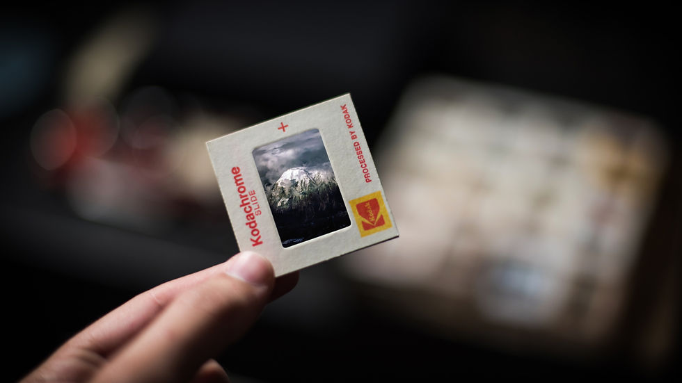 Hand holding a Kodachrome slide with a mountain scene. Text reads "Kodachrome Slide" and "Processed by Kodak." Blurred background.