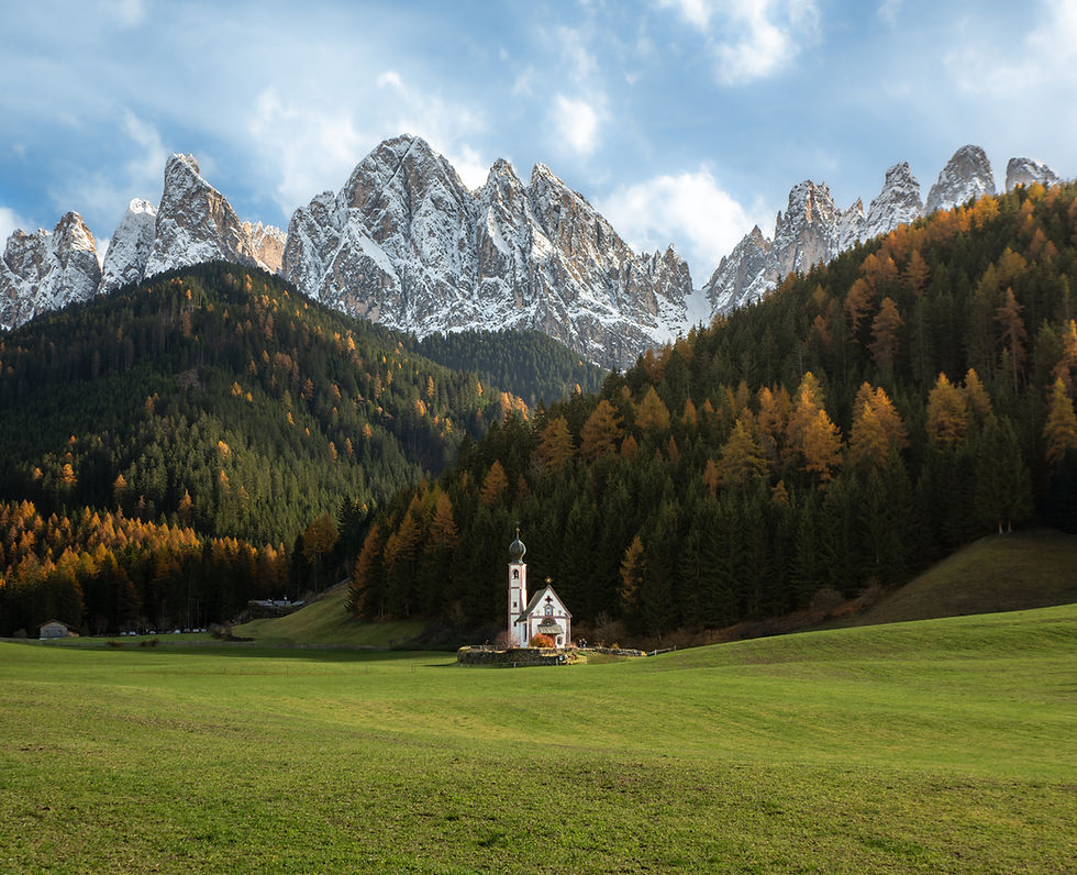 San Giovanni Church in Val di Funes in the Dolomites with green meadow, tree-covered hills, and snow-capped mountains in the background