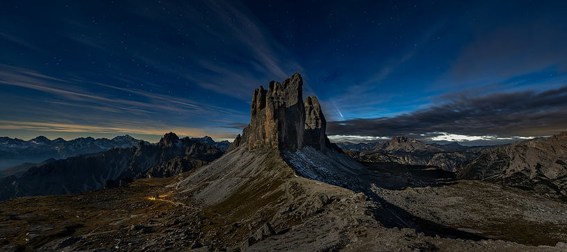 The peaks of Tre Cime di Lavaredo in the Dolomites, taken at night and illuminated by the moon. The sky is dark blue and there is a comet in the sky above the peak.