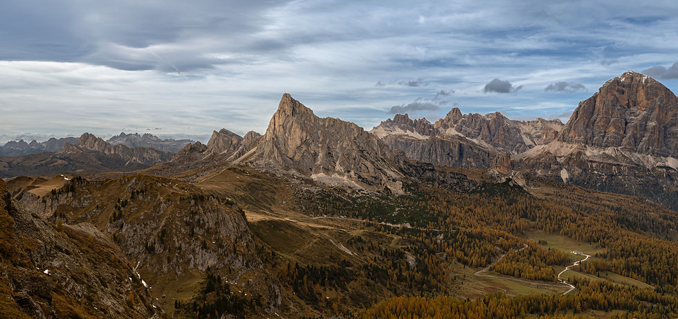 The mountains around Passa di Giau in the Dolomites under a cloudy sky, with rugged peaks and a forested valley below. Browns and greens dominate the landscape.