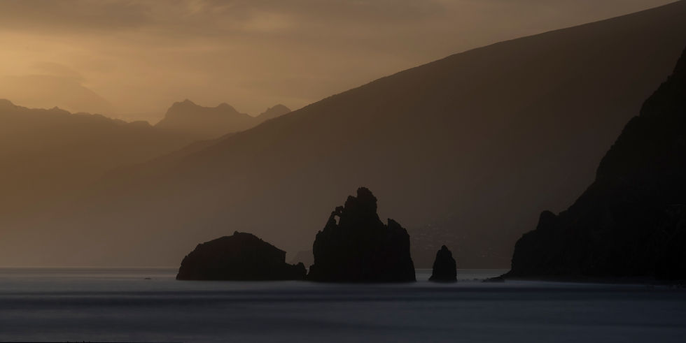 The sea stacks of Ribeira da Janela seen from Porto Moniz, silhouetted against layered mountains and an orange sky.