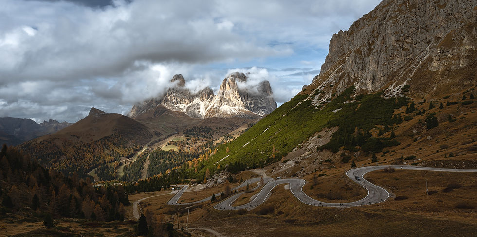 Winding road leads through a mountainous landscape with the dramatic peaks of Sassolunga shrouded in clouds. Autumn foliage adds warm tones to this scene from the Dolomites.