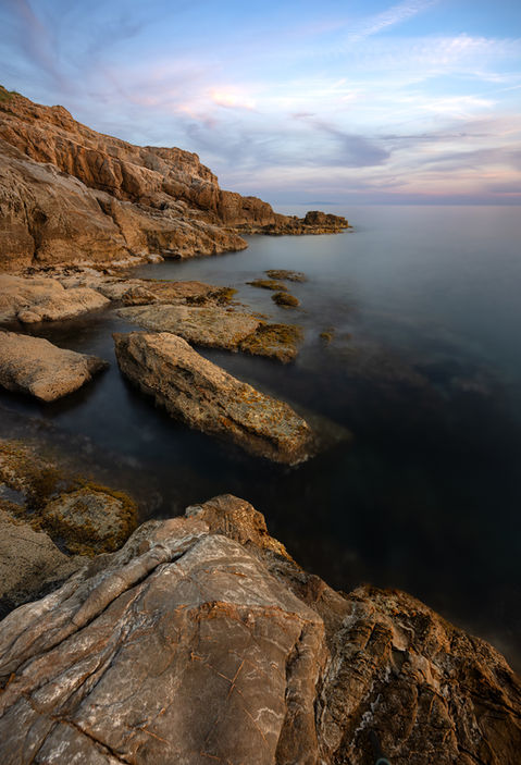 A coastal scene of rock formations with smooth water and pastel colours in the sky, taken on the coast south of Livorno