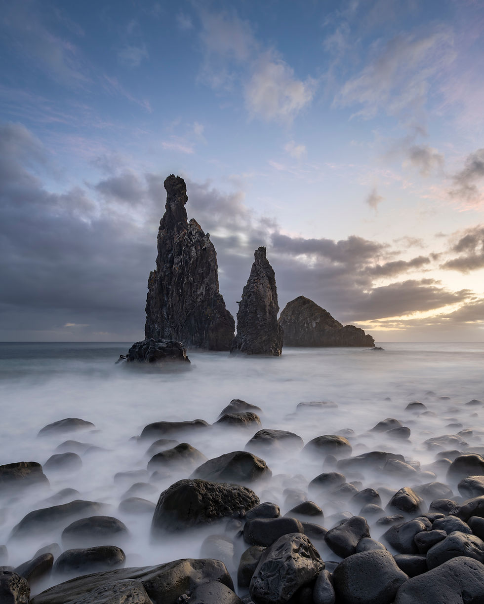 The sea stacks at Ribeira da Janela in Madeira at sunrise, with soft water flowing over pebbles beneath a blue and yellow sky.