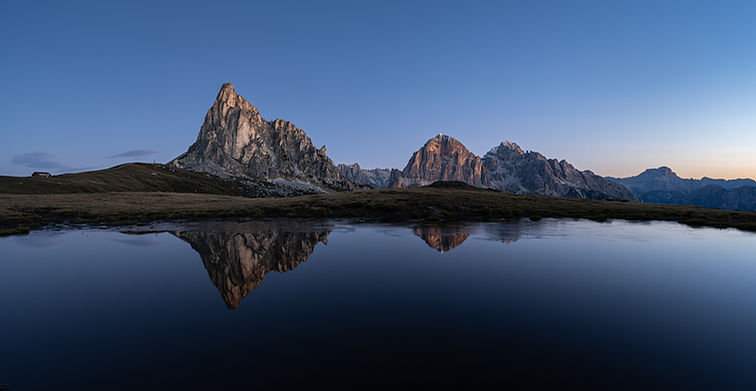 The peak of Passa di Giau with alpenglow from the sunrise, reflected in a still pond under a clear blue sky.