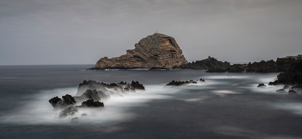 Long exposure of the sea at Porto Moniz in Madeira, with water blurring over rocks and a sea stack beneath a grey sky.