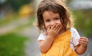 a little girl with light brown hair, outside, wearing a white shirt and orange jumpsuit, covering her smile behind her hand