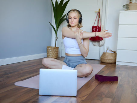 A woman sits cross-legged on a yoga mat in a home setting, gently stretching her arm while following a guided somatic exercise session on her laptop, with a resistance band placed beside her.
