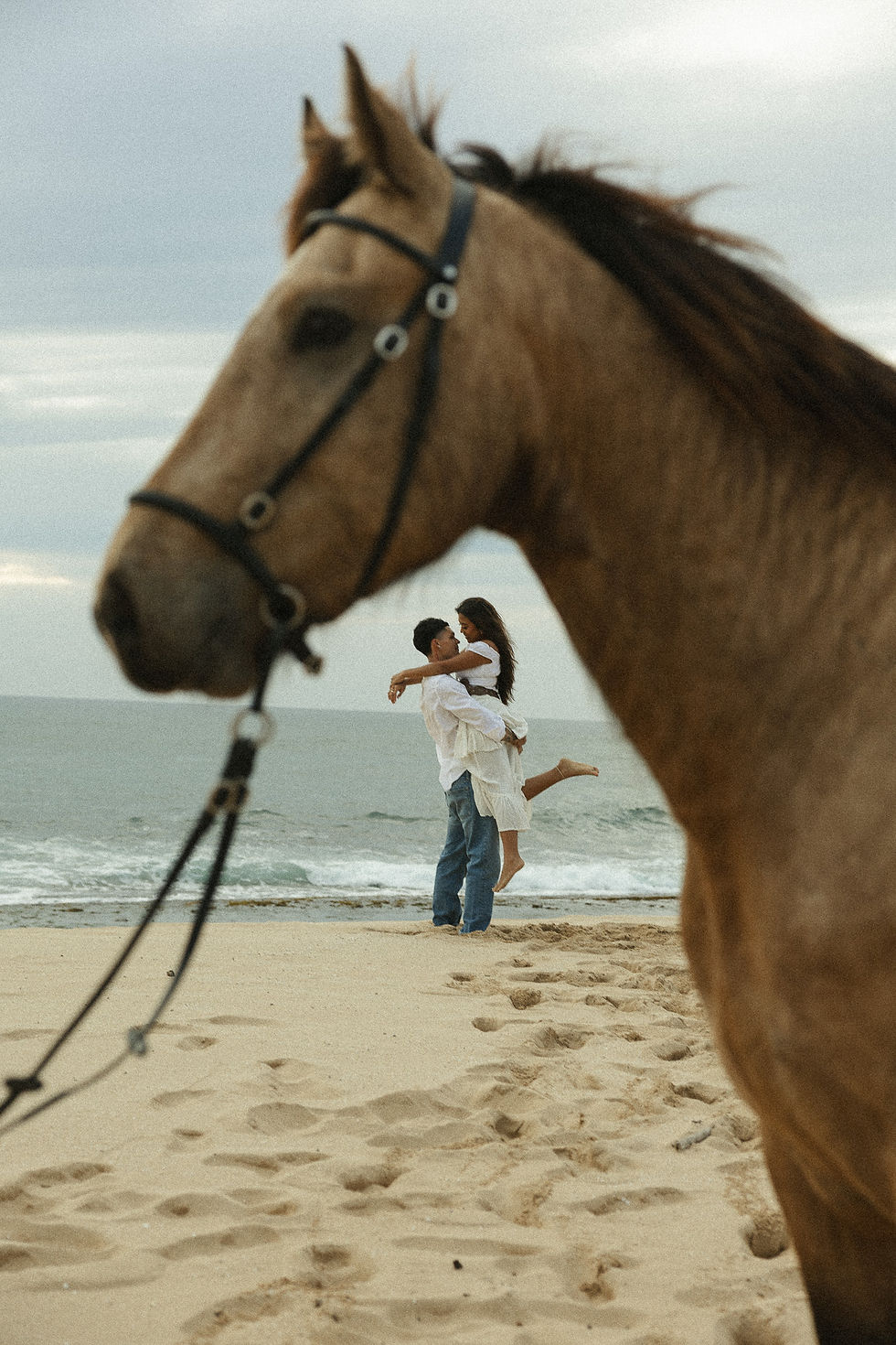 Couple embracing on a beach framed by a horse in the foreground.