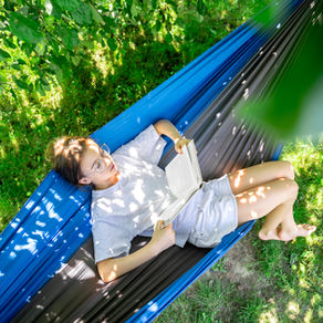 Dospívající dívka relaxuje v houpací síti na zahradě a čte si knihu. Fotografie ilustruje význam odpočinku, zpomalení a regenerace během letních prázdnin po náročném školním roce a podporu zdravého psychického vývoje dospívajících.