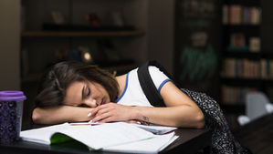 Dospívající dívka usnula nad otevřenými sešity ve škole či knihovně, což ilustruje nedostatek spánku a změny spánkového rytmu typické pro období adolescence.