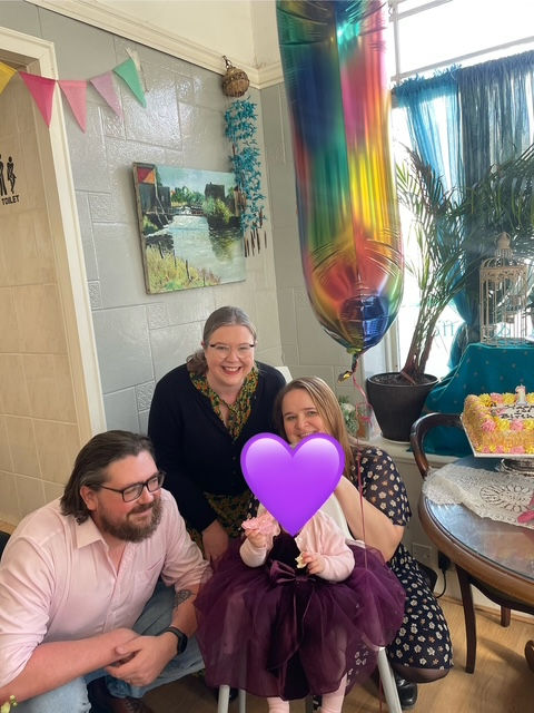 London celebrant Mair Garland is posing with two parents and a baby at a humanist naming ceremony at All Things Nice cafe, Ewell. There is a rainbow '1' balloon. The baby's face is obscured with a purple heart. There is a pink and yellow cake on the table.