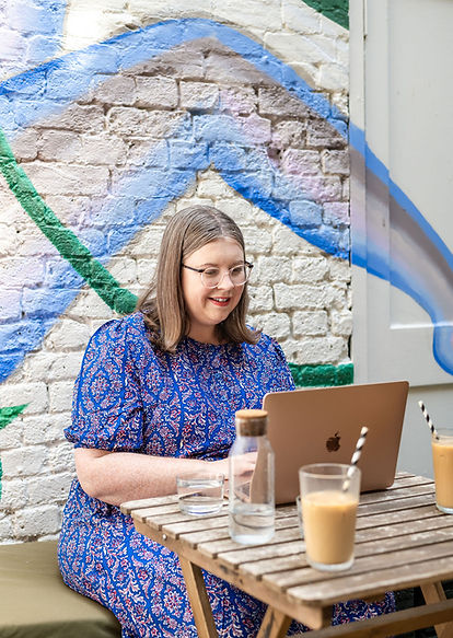 London celebrant Mair Garland is sitting at her laptop at Spinach cafe in Crystal Palace. The white bricked wall behind her has green and blue grafitti and there are two iced lattes an water on the table.