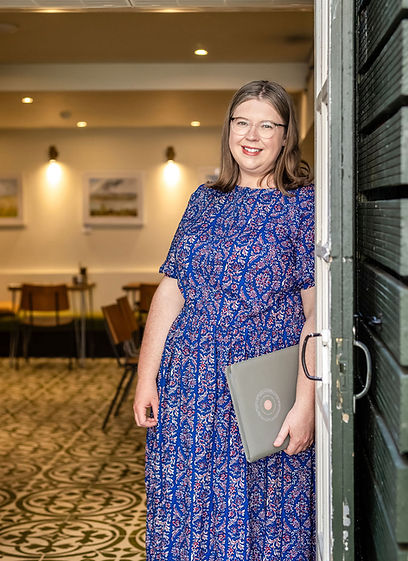 London celebrant Mair Garland standing in the doorway of Spinach restaurant, Crystal Palace in south-east London. Mair is smiling and is wearing a blue dress with a red paisley pattern and is holding her grey ceremony foder with the 'Garland Ceremonies' logo on the front.