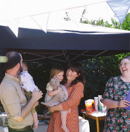 London celebrant Mair Garland leads a naming ceremony for two children in a garden ceremony. Mair, the children and the parents are laughing. There's a marquee and bunting in the background. 