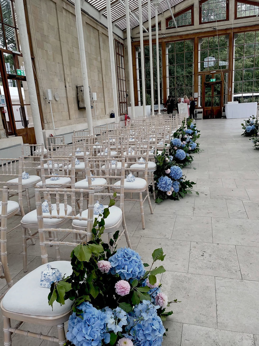 Wedding at The Nash Conservatory Kew Gardens, led by London wedding celebrant Mair Garland. There are rows of chairs, each with a blue and white paper teapot favour bag adorned with blue hydrangeas and pink dahlias at the end of each row. 