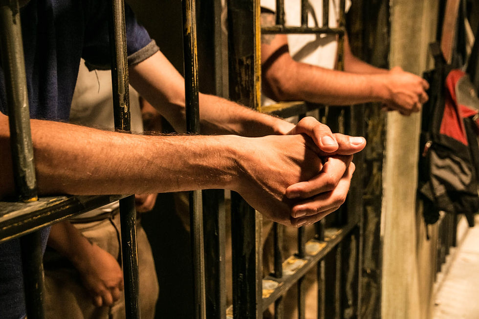 Hands gripping prison bars in a dimly lit cell, with tense emotions. Beige and blue clothing visible in the background.
