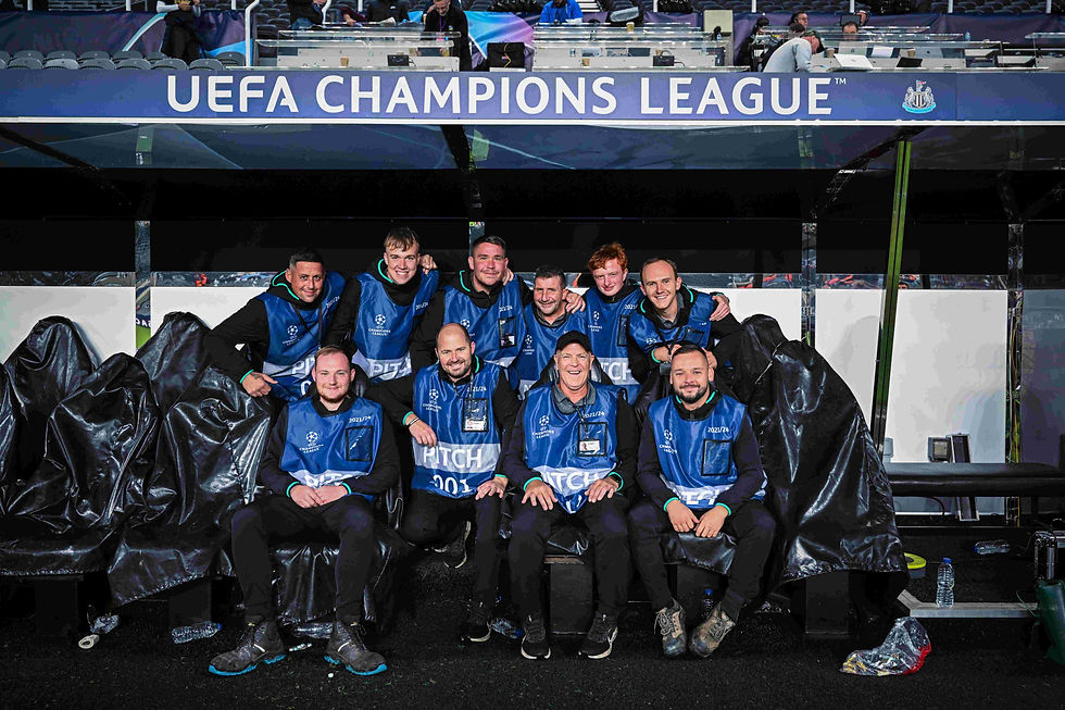 Ten smiling people in blue vests pose under a "UEFA Champions League" sign on a stadium bench. Joyful mood, dimly lit setting.