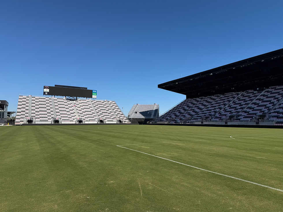 Empty soccer stadium with green pitch, patterned seats, blank scoreboard, and a clear blue sky.