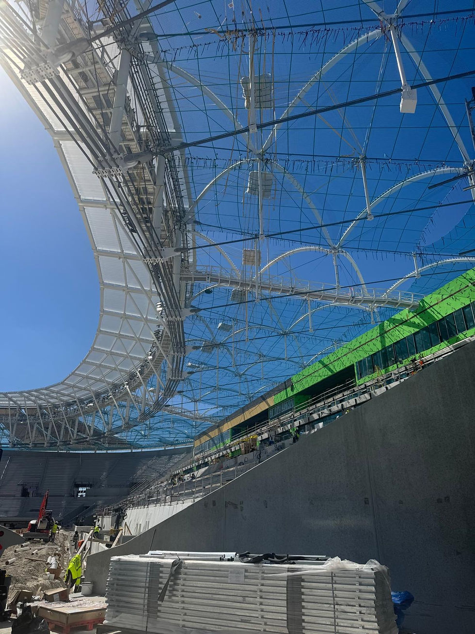 Workers build a stadium under a blue canopy and white steel roof, with green facade panels and stacked materials in sunlight.