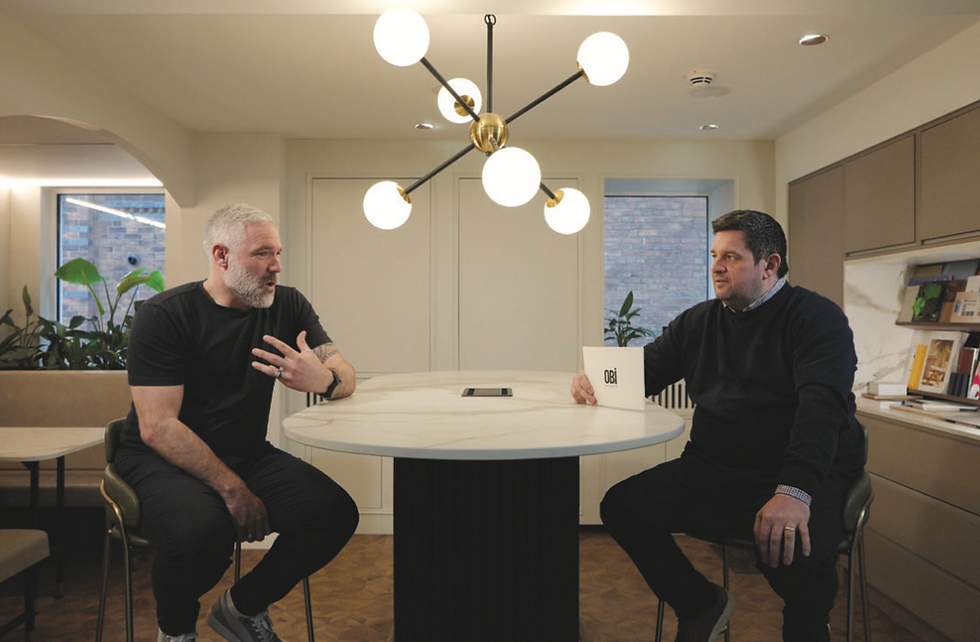 Two men sit talking at a round table in a modern office with plants. One holds a mug labeled "Obi." Bright light fixture overhead.
