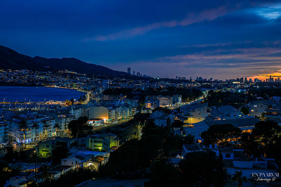 Cityscape at dusk with illuminated buildings and roads. Mountains in the background, sky in deep blue and orange hues over the horizon.